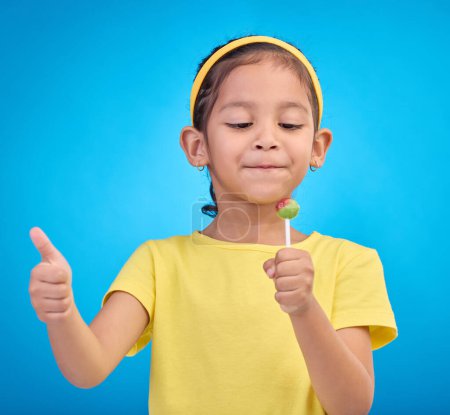 Candy, happy and child with lollipop and thumbs up on blue background with happiness, smile and excited. Party, childhood and young girl with hand sign eating sweets snack, sugar and treats in studio.