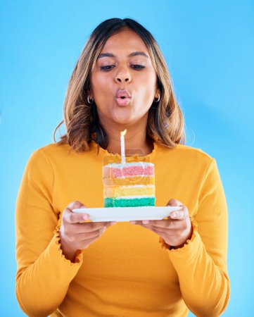 Birthday cake, woman and blow candles in studio, blue background and celebration. Female model, dessert and wishing at party, flame and special event, rainbow treat or celebrate happiness on backdrop.