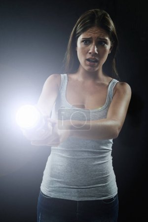 Photo for Whos out there. A young brunette holding a flashlight while looking scared and isolated on a black background - Royalty Free Image