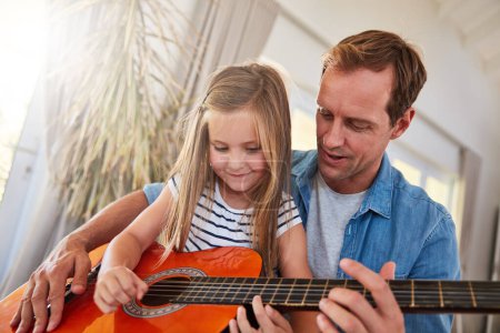 Youre catching on quick. a father and his young daughter sitting together in the living room at home playing guitar