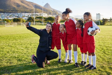 Fußball, Mannschafts- und Trainer-Selfie mit dem Handy auf dem Spielfeld nach dem Training, Training oder Spiel in einem Sportverein. Fußball-Mädchengruppe lächelt und freut sich über gemeinsames Foto für soziale Medien auf Sportplatz.