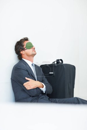 Foto de Hombre de negocios con máscara para dormir. Hombre de negocios maduro con una máscara para dormir sentado contra la pared con equipaje de viaje - Imagen libre de derechos