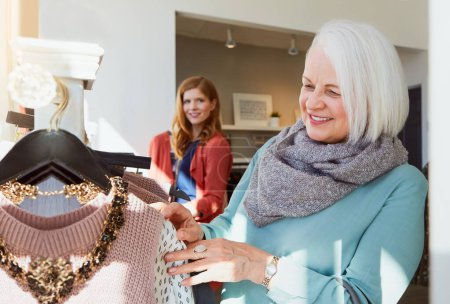 Foto de La gente admira su estilo. una mujer mayor comprando en una boutique de ropa con una mujer mirándola al fondo - Imagen libre de derechos