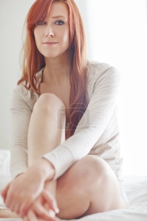 Téléchargez les photos : Portrait de belle rousse assise seule sur un lit à la maison. Un modèle de tête rouge se sentant flirté et sexy tout en posant séduisante dans une chambre le week-end. Jeune femme relaxante et sensuelle. - en image libre de droit