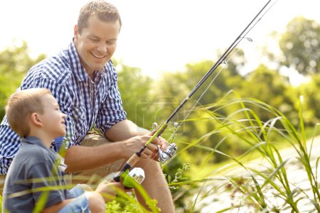 The fish are on their way, son. Father and son sitting together beside a lake with their fishing rods
