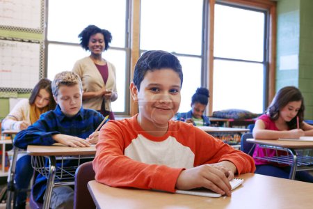 Téléchargez les photos : Nous apprenons pour la vie pas seulement pour un test. un jeune garçon assis en classe avec son professeur et ses camarades de classe brouillés en arrière-plan - en image libre de droit