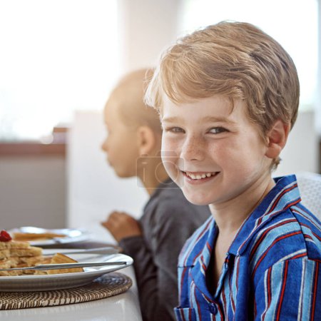 Téléchargez les photos : C'est l'heure du petit déjeuner. un petit garçon déjeunant avec sa sœur floue en arrière-plan - en image libre de droit