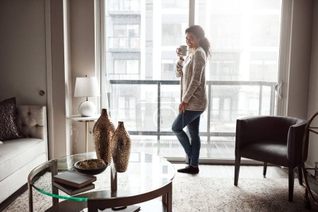 Téléchargez les photos : Pourquoi sortir quand tu peux rester à la maison ? une jeune femme joyeuse buvant du café en regardant par une fenêtre à l'intérieur de la maison pendant la journée - en image libre de droit