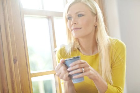 Photo for A beautiful woman smiling and sitting near a window inside a house. Happy attractive blonde woman chilling drinking coffee. Happy female relaxing and daydreaming at home off the grid in a sunroom. - Royalty Free Image