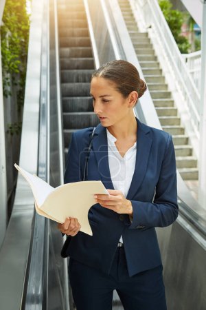 Téléchargez les photos : Je me prépare pour sa prochaine réunion. une jeune femme d'affaires lisant des documents sur un escalier roulant - en image libre de droit