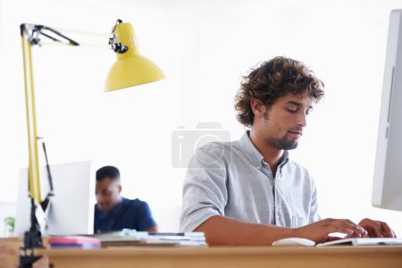 Foto de Tengo que cumplir el plazo... un joven hombre de negocios guapo sentado en su escritorio con su colega en el fondo - Imagen libre de derechos