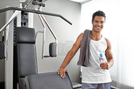 Foto de Tengo confianza en mi salud y estado físico. Un joven étnico de pie junto a una máquina de ejercicios en un gimnasio - Imagen libre de derechos