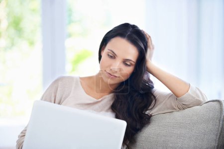 Photo for Surfing the web at home. A young woman sitting on her sofa and looking at her laptop screen while relaxing - Royalty Free Image