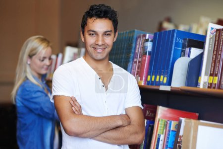 Foto de Se siente seguro de la universidad. Retrato de un joven estudiante guapo de pie con confianza en la biblioteca - Imagen libre de derechos