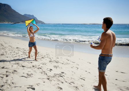 Téléchargez les photos : Voyons jusqu'où on peut aller. Un jeune couple amusant jouant avec un cerf-volant tout en profitant d'une journée sur la plage - en image libre de droit