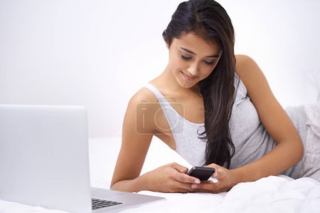 Téléchargez les photos : Rester sociable. Une jeune femme au lit avec son téléphone et son ordinateur portable - en image libre de droit