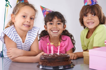 Foto de Haciendo recuerdos de infancia. Tres amigos en una fiesta de cumpleaños sonriendo con un pastel delante de ellos - Imagen libre de derechos