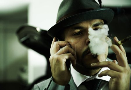 Foto de Violencia en sus ojos. Un mafioso arrogante soplando humo en el aire mientras habla por su celular - Imagen libre de derechos
