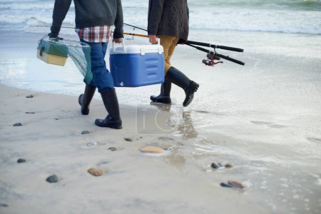 Foto de Herramientas del comercio pesquero. Dos pescadores llevando un refrigerador y una caja de aparejos en la playa por la mañana temprano - Imagen libre de derechos