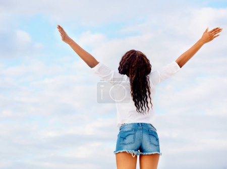 Foto de Abrazando su vida. Vista trasera de una joven mujer de pie contra un cielo azul con los brazos extendidos - Imagen libre de derechos