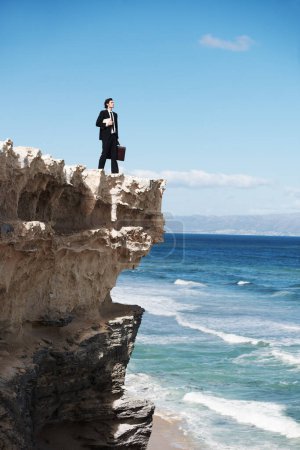 Photo for I can conquer the world. Young businessman holding a briefcase and a book standing on the edge of a cliff overlooking the ocean - Royalty Free Image