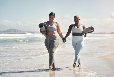 Photo for We dont catch up over coffee, we catch up over yoga. two young women walking on the beach with their yoga mats - Royalty Free Image