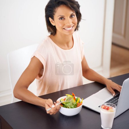 Téléchargez les photos : Déjeuner satisfaisant et sain à la maison. Portrait d'une jeune femme dégustant une salade et travaillant sur un ordinateur portable à la maison - en image libre de droit