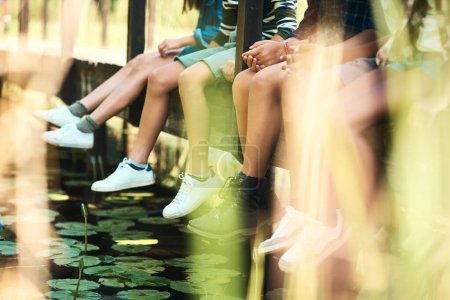 Foto de La naturaleza sabe cómo hacer que los niños se relajen. un grupo de adolescentes irreconocibles sentados en un puente en la naturaleza en el campamento de verano - Imagen libre de derechos