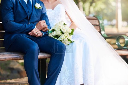 Photo for Closeup of a bride and groom sitting on a park bench together. Man wearing suit and woman wearing wedding dress and holding bouquet while sitting outdoors. - Royalty Free Image