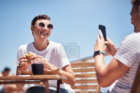 Foto de Quédate quieto mientras tomas una foto. una joven alegre tomando su foto por su novio afuera mientras almorzan - Imagen libre de derechos