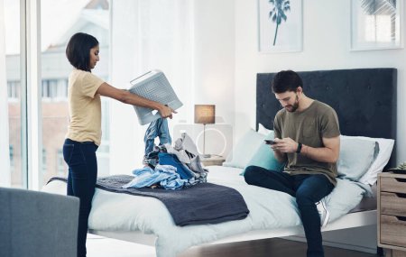 Photo for Whos doing the lions share of the housework. a young woman doing laundry while her husband uses his smartphone at home - Royalty Free Image