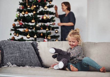 Téléchargez les photos : Je m'occupe. une adorable petite fille assise sur le canapé pendant que sa mère fixe le sapin de Noël derrière elle - en image libre de droit