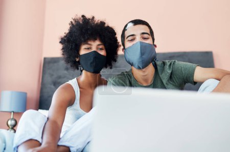 Téléchargez les photos : Restons ici et sauvons des vies. un jeune couple portant des masques lorsqu'il utilise un ordinateur portable à la maison - en image libre de droit