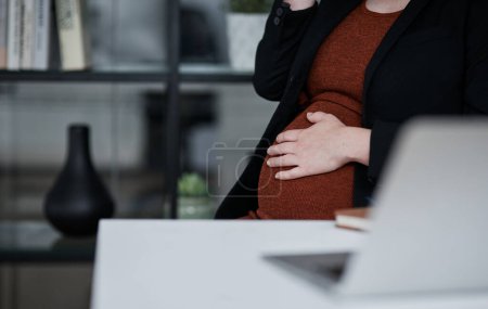 Téléchargez les photos : Mes jours au bureau raccourcissent. une femme d'affaires méconnaissable assise seule au bureau et tenant son estomac enceinte - en image libre de droit