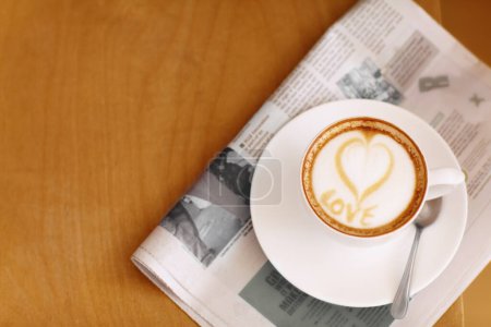 Téléchargez les photos : Café, cappuccino et coeur en mousse avec journal sur une table sans personne dans un restaurant. Café boisson, écriture d'amour et l'art de la boisson mousseuse avec du lait dans un café avec un gros plan de tasse et de maquette. - en image libre de droit