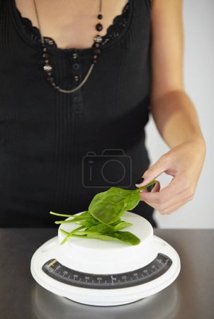 Photo for Hand, scale and spinach in kitchen for diet, food or eating disorder with leaves for meal plan. Woman, mental health and vegetable leaf to lose weight with anorexia, bulimia or starving in home. - Royalty Free Image
