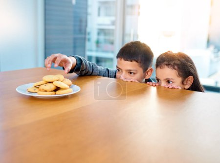 Foto de Voy a entrar, cúbreme hermana. dos niños traviesos robando galletas en la mesa de la cocina en casa - Imagen libre de derechos