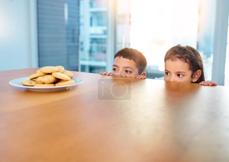Foto de Tenemos que atacar rápido y no dejar rastros. dos niños traviesos robando galletas en la mesa de la cocina en casa - Imagen libre de derechos