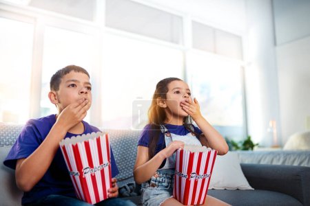 Foto de Acción de apertura de boca y entretenimiento. dos niños pequeños sentados en un sofá y comiendo palomitas de maíz mientras ven películas en casa - Imagen libre de derechos