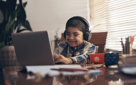 Téléchargez les photos : L'école la plus cool. un adorable petit garçon utilisant un ordinateur portable et un casque tout en effectuant une affectation scolaire à la maison - en image libre de droit