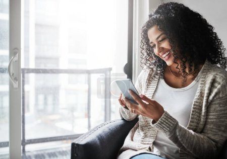 Téléchargez les photos : Rester connecté. une jeune femme joyeuse se détendre tout en envoyant des SMS sur son téléphone portable à la maison pendant la journée - en image libre de droit