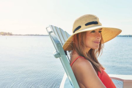 Photo for This is my home. a cheerful young woman wearing a hat while being seated on a chair next to a lake outside in the sun - Royalty Free Image