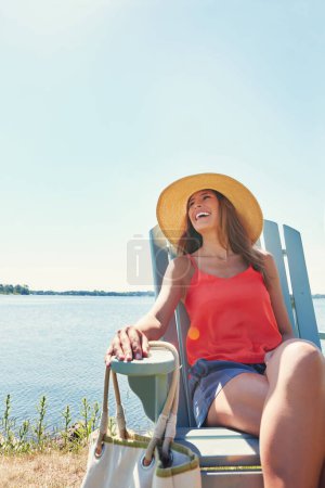 Téléchargez les photos : Rien et personne ne peut gâcher son humeur. une jeune femme joyeuse portant un chapeau tout en étant assis sur une chaise à côté d'un lac dehors au soleil - en image libre de droit