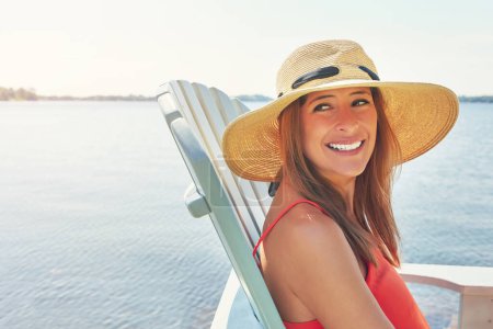 Téléchargez les photos : Un des endroits les plus paisibles. une jeune femme joyeuse portant un chapeau tout en étant assis sur une chaise à côté d'un lac dehors au soleil - en image libre de droit