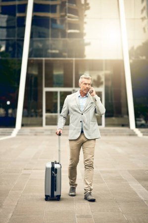 Foto de Acabo de aterrizar hace unos minutos. un hombre de negocios maduro hablando por celular mientras camina con una maleta en la ciudad - Imagen libre de derechos