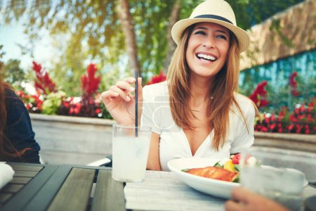 Foto de ¿Cómo está tu bebida? una joven alegre vistiendo un sombrero y disfrutando de una bebida fría mientras se sienta en un restaurante al aire libre durante el día - Imagen libre de derechos