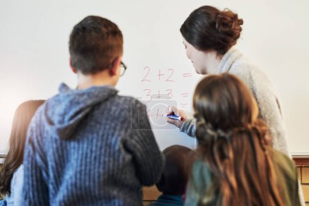 Photo for Who knows the answer to this one class. Rearview shot of teacher and her students doing math equations on a white board in class - Royalty Free Image