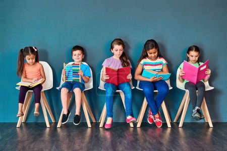 Foto de Theres a lot to learn. Studio shot of a group of kids sitting on chairs and reading books against a blue background - Imagen libre de derechos
