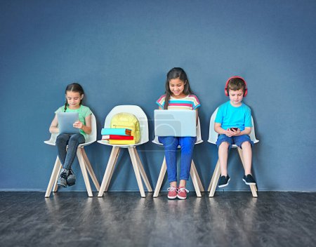 Téléchargez les photos : Growing up in a wireless world. Studio shot of kids sitting on chairs and using wireless technology against a blue background - en image libre de droit