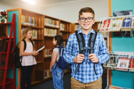 Photo for Geared up and ready for learning. Portrait of a cheerful young boy wearing a schoolbag while standing inside of library during the day - Royalty Free Image